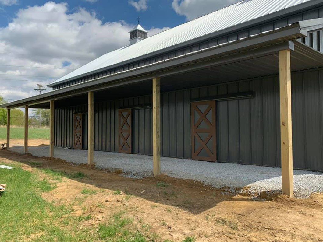Gray metal barn with a covered porch supported by wooden posts. Three brown sliding doors are visible.