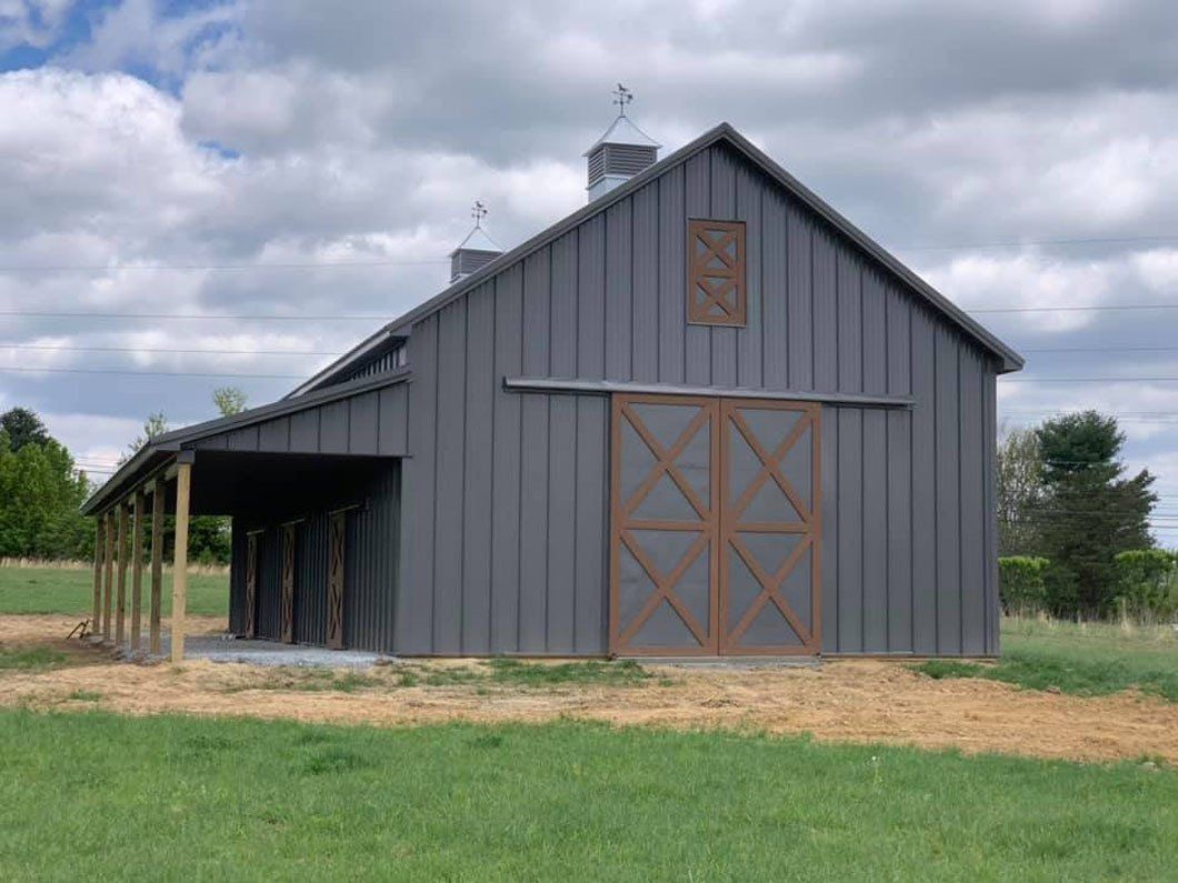 Gray barn with a brown sliding door, set in a green field under a cloudy sky. There is a covered porch attached to the side.