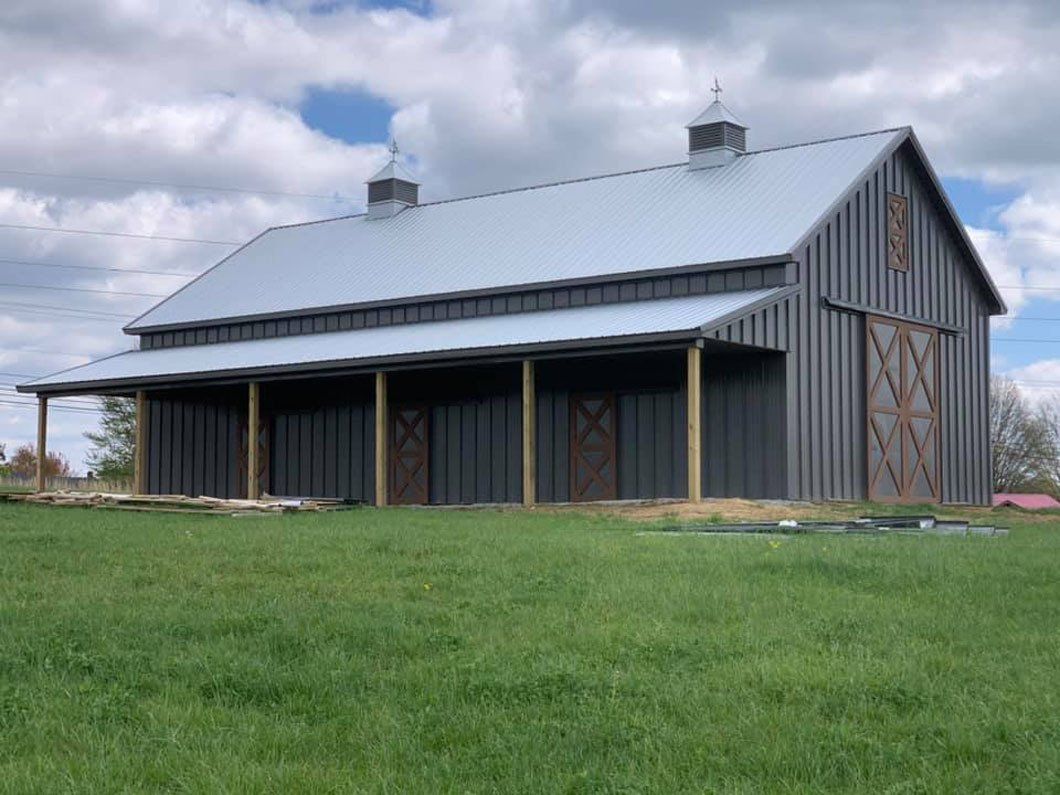 Gray barn with a silver roof and covered porch, set in a grassy field under a cloudy sky.