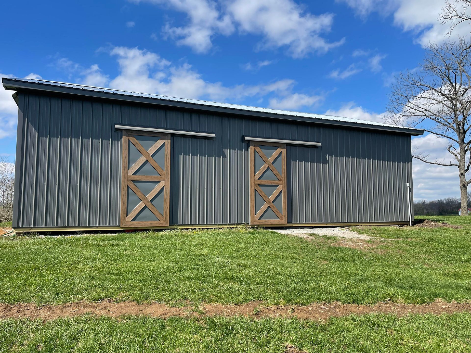 Gray metal shed with two sliding wooden doors, set in a grassy field under a partly cloudy blue sky.