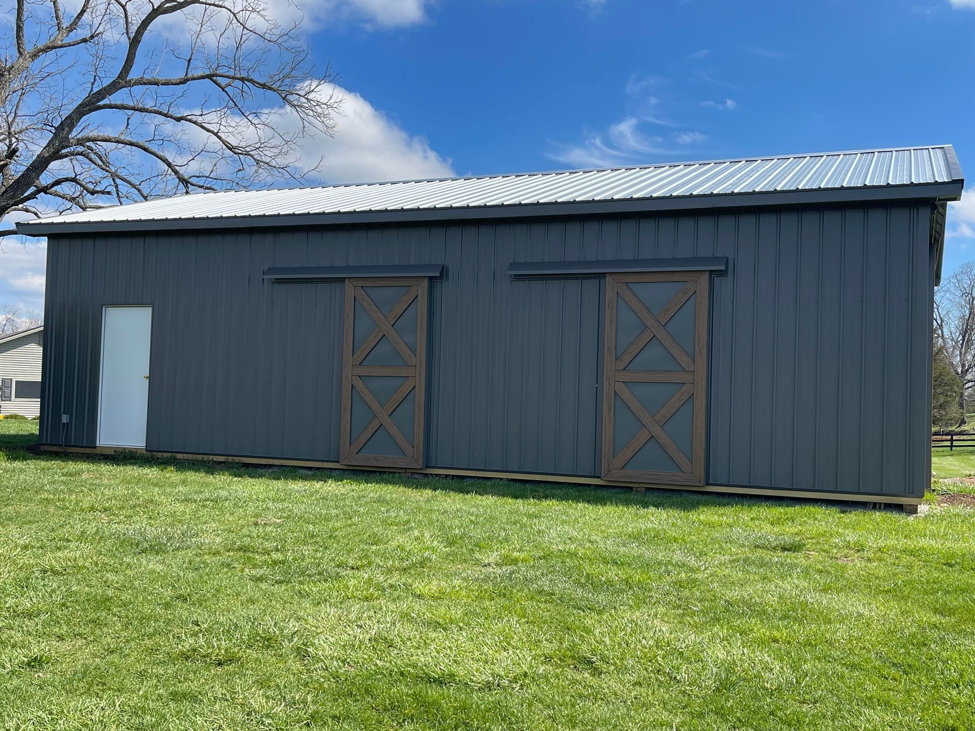 Gray barn with a metal roof and wooden sliding doors, set on a grassy field under a blue sky.