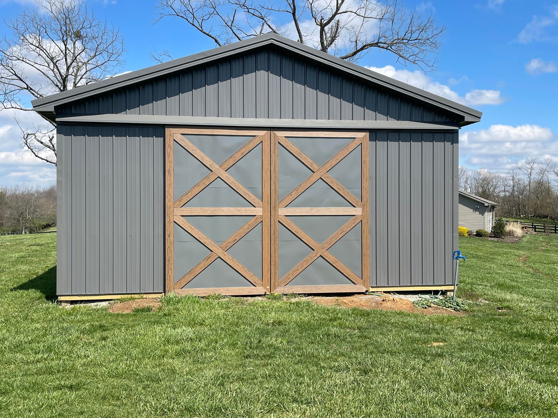 Gray shed with wooden barn doors in a grassy field under a blue sky.