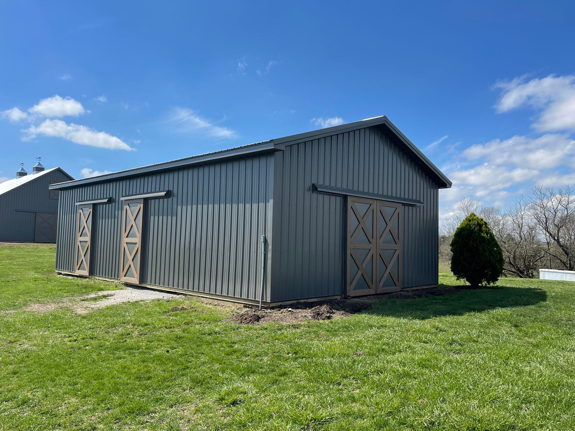Gray metal barn with sliding wooden doors on a grassy field under a blue sky. Another building is in the background.