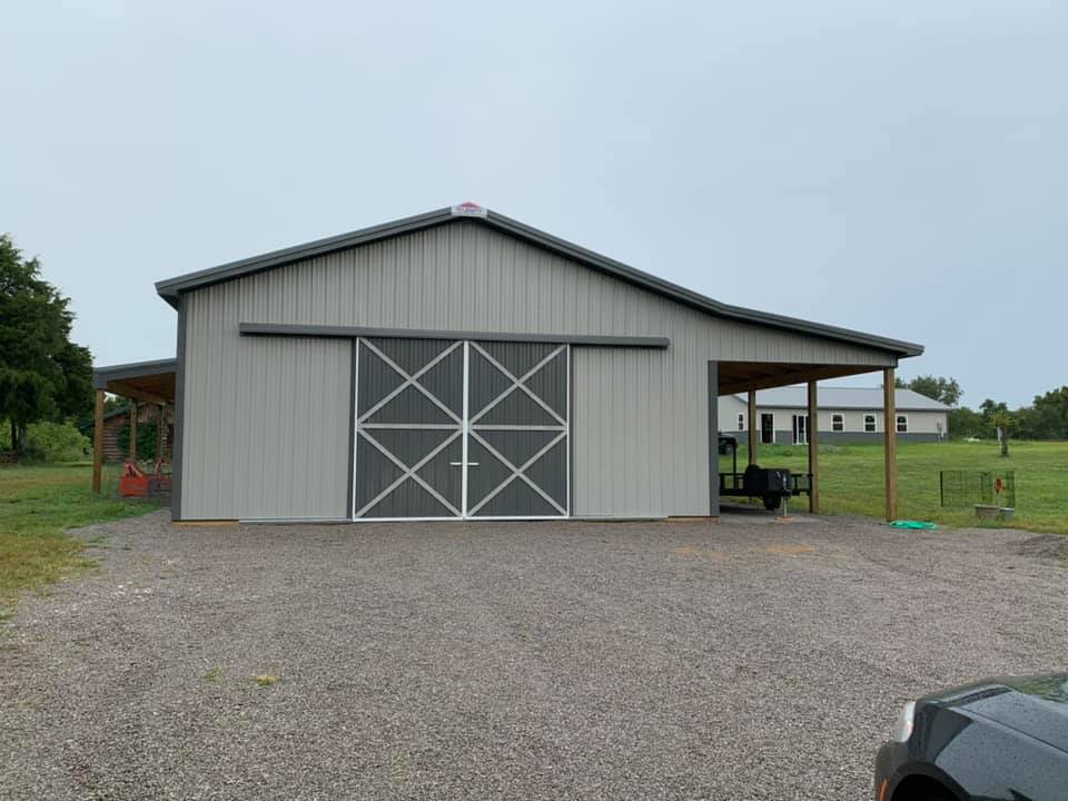 Gray metal barn with large sliding doors, a gravel driveway, and a covered side area. Cloudy sky.