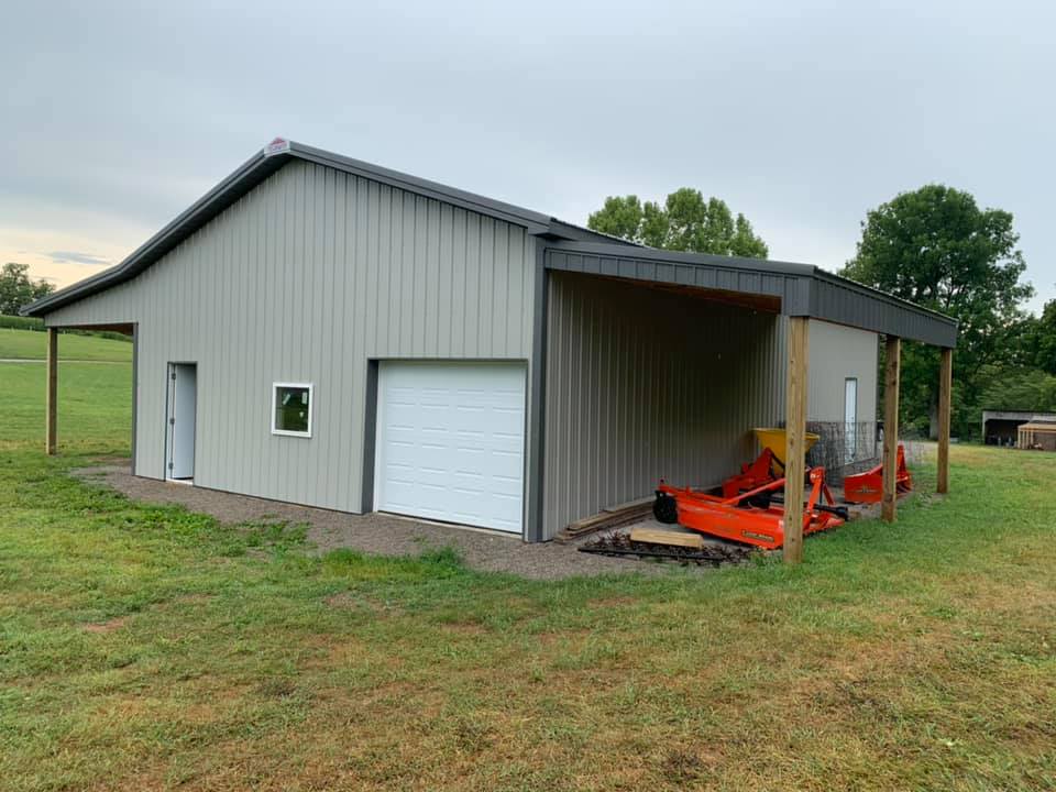 A tan metal barn with an attached carport on a grassy hill. A white garage door and small window are visible on the front.