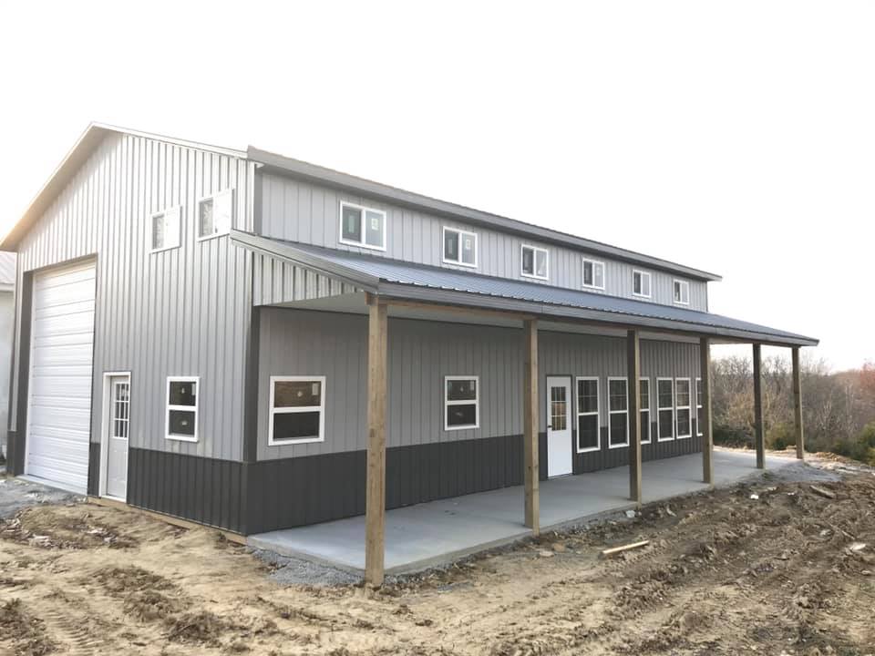 A large, two-tone gray metal building with a porch. The building has a garage door, windows, and a concrete porch supported by wooden pillars.