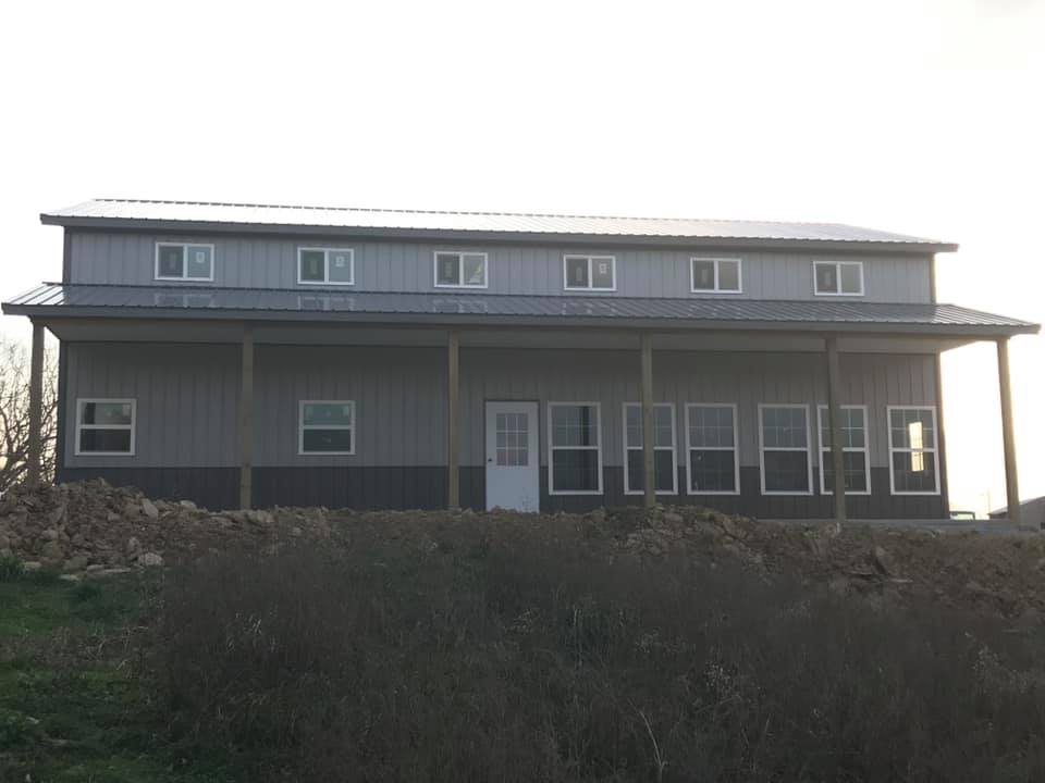 Two-story gray metal building with a porch and multiple windows. The building sits on a slight hill, against a muted sky.