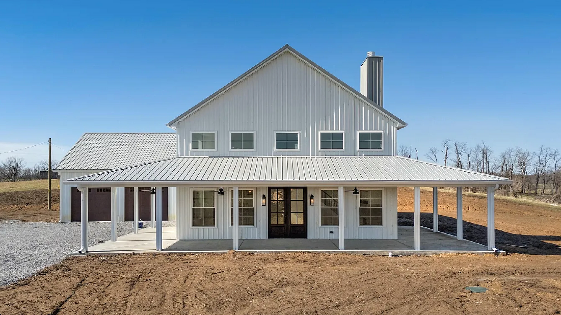 White farmhouse with a covered porch and metal roof, set against a blue sky in a rural area.