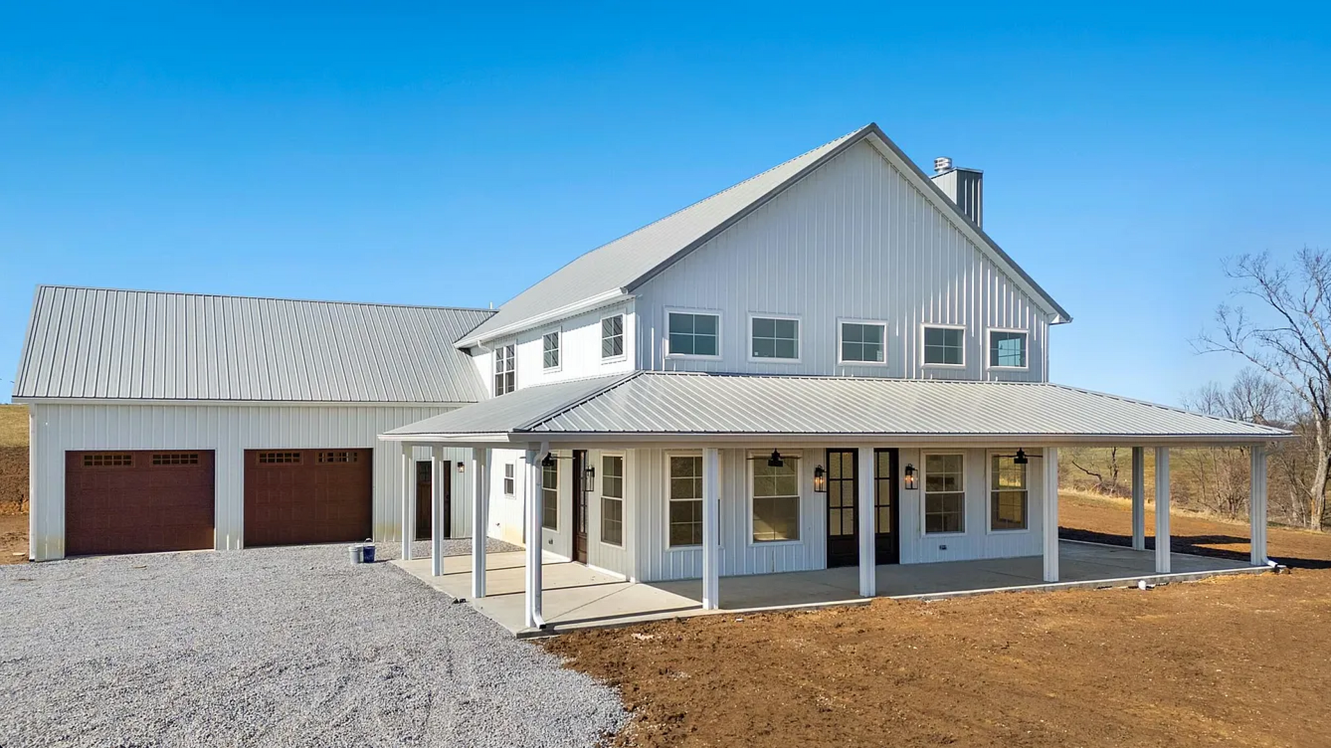 Farmhouse with white siding, a metal roof, and a wraparound porch. There's a two-car garage on the left, and the house is set against a blue sky.