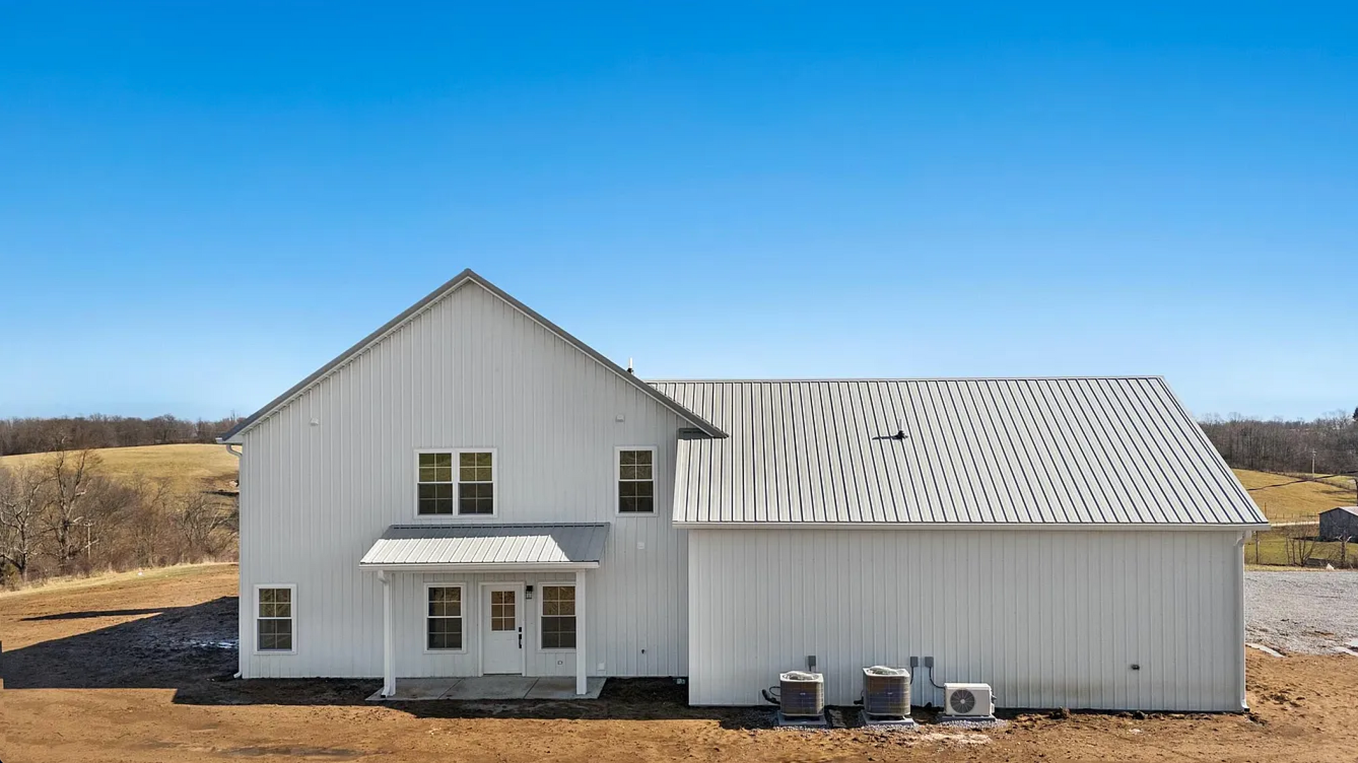 White farmhouse with a gable roof and a lower, attached structure against a blue sky. Two chairs and an AC unit sit on the ground.