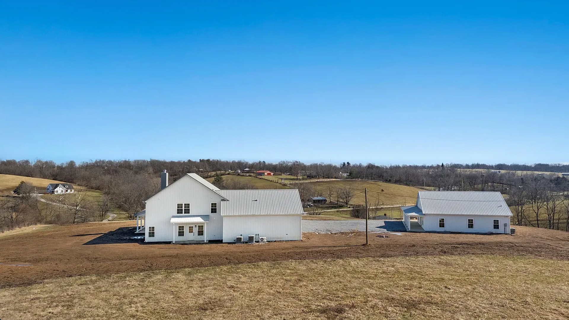 White farmhouse and detached building on a brown field under a blue sky, surrounded by trees.