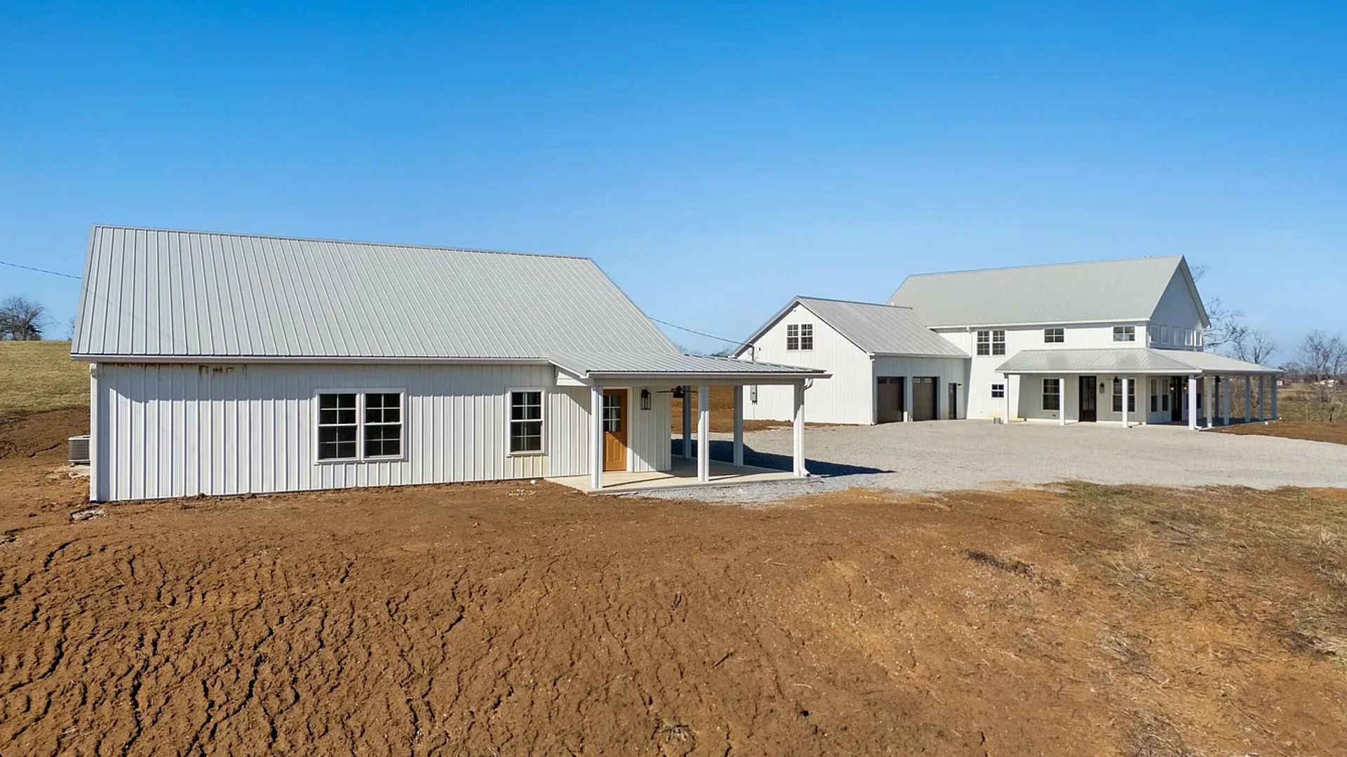 White farmhouse under construction on a hillside. Features a barn-style structure and a larger house with a porch, under a clear blue sky.