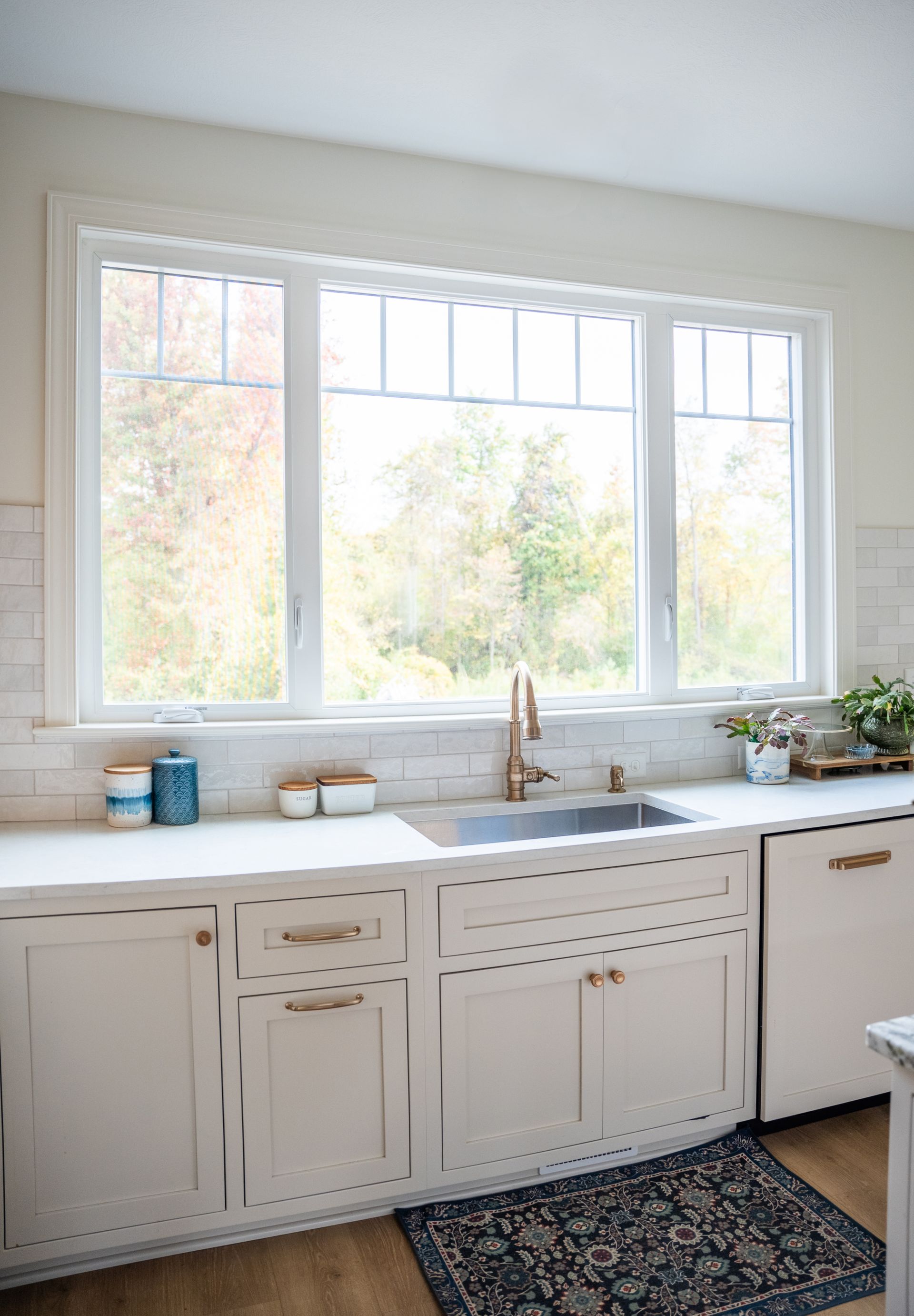 Cream-colored kitchen cabinets with a white countertop, sink, and gold faucet in front of a large window with a scenic view.