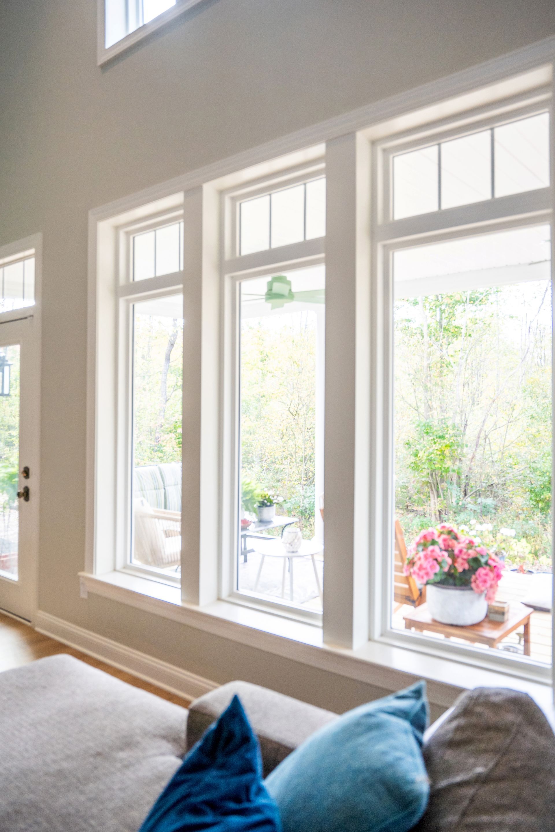 Living room with three tall windows overlooking a backyard patio. White trim frames the windows and doorway. A gray couch with blue pillows is in the foreground.