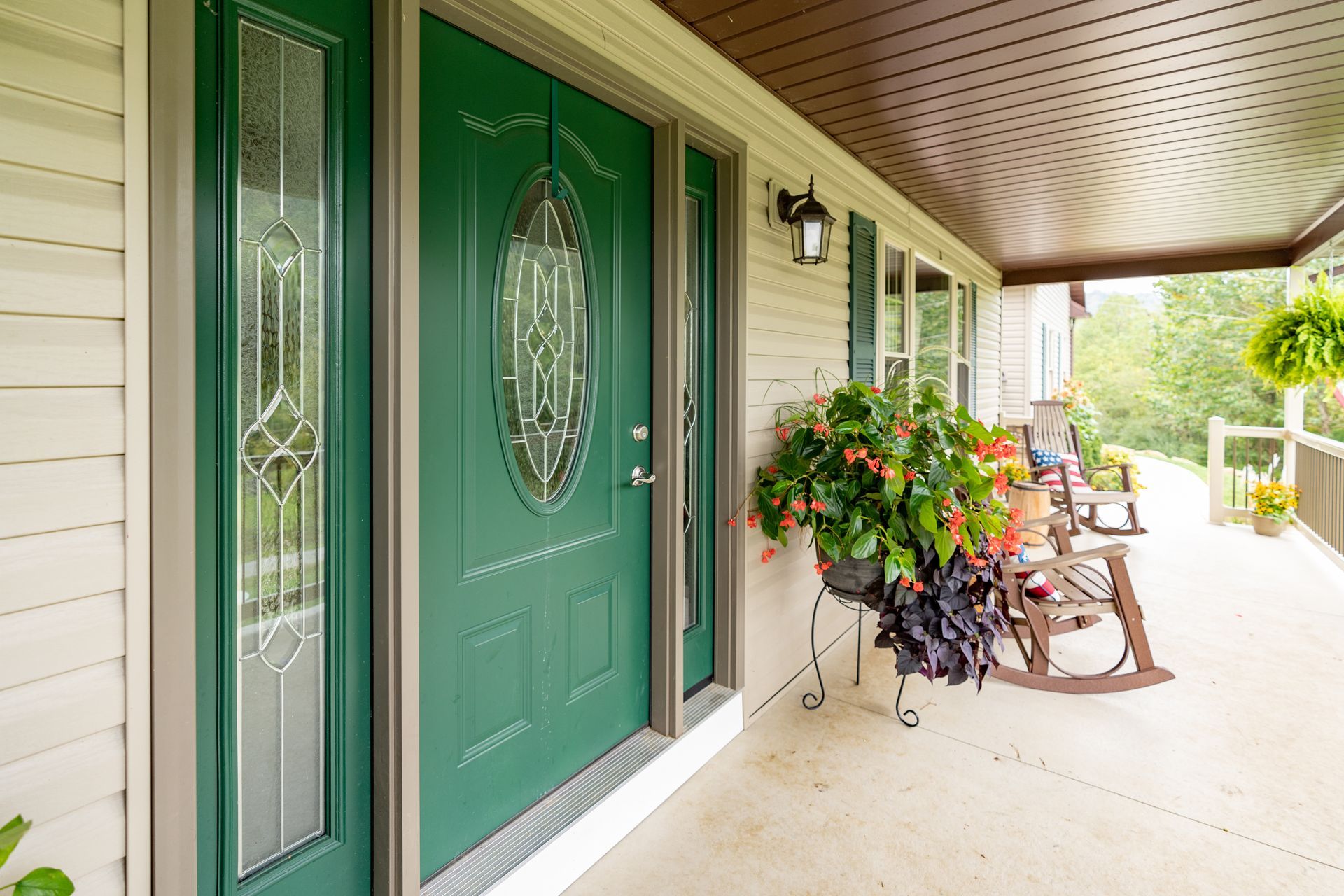 Green front door with sidelight and porch with rocking chairs and potted plants.