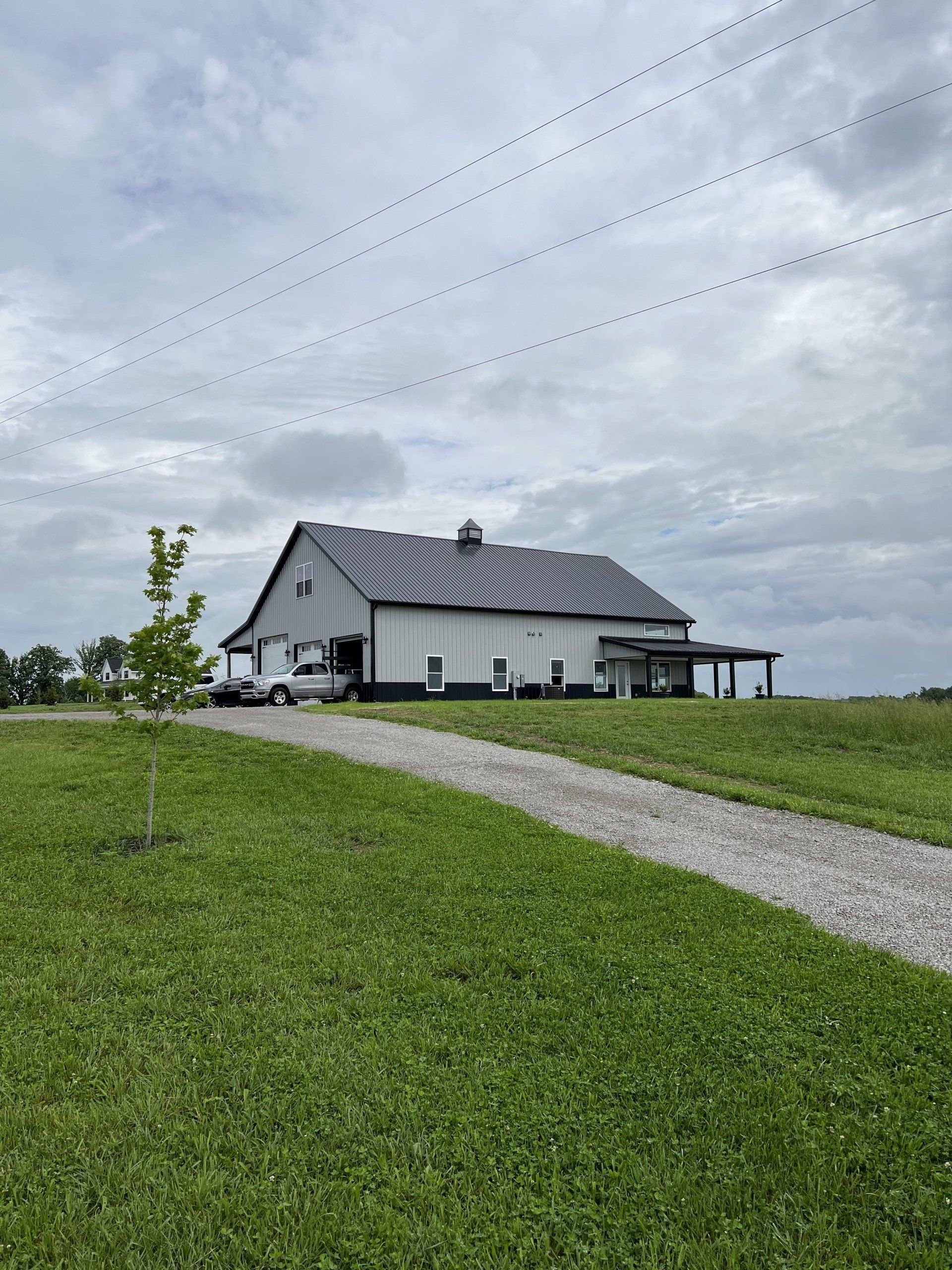 A white barn with a black roof sits on a grassy hill under a cloudy sky. A gravel driveway leads up to the barn.