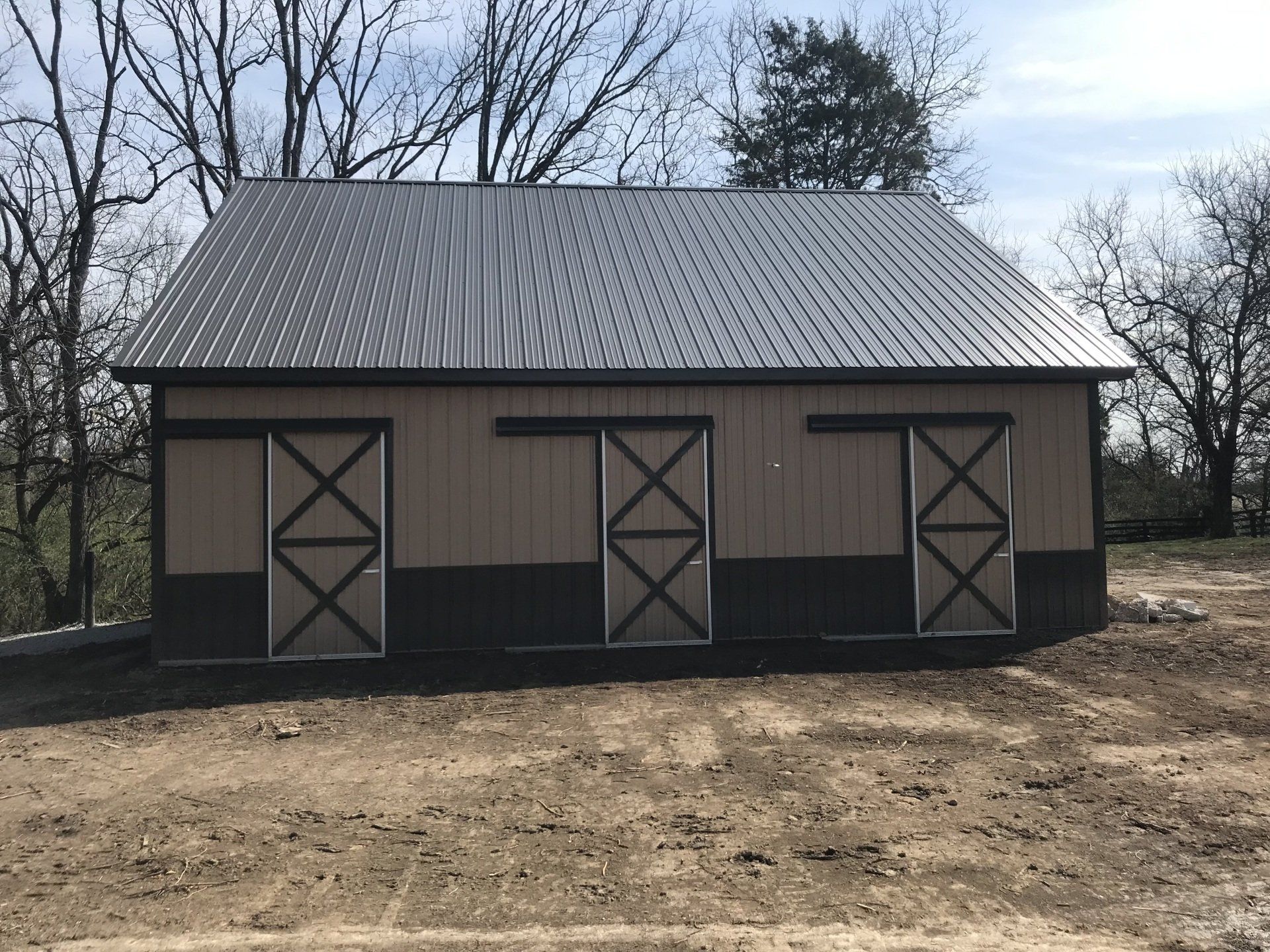 Tan and brown barn with three sliding doors, a gray metal roof, and bare trees in the background.