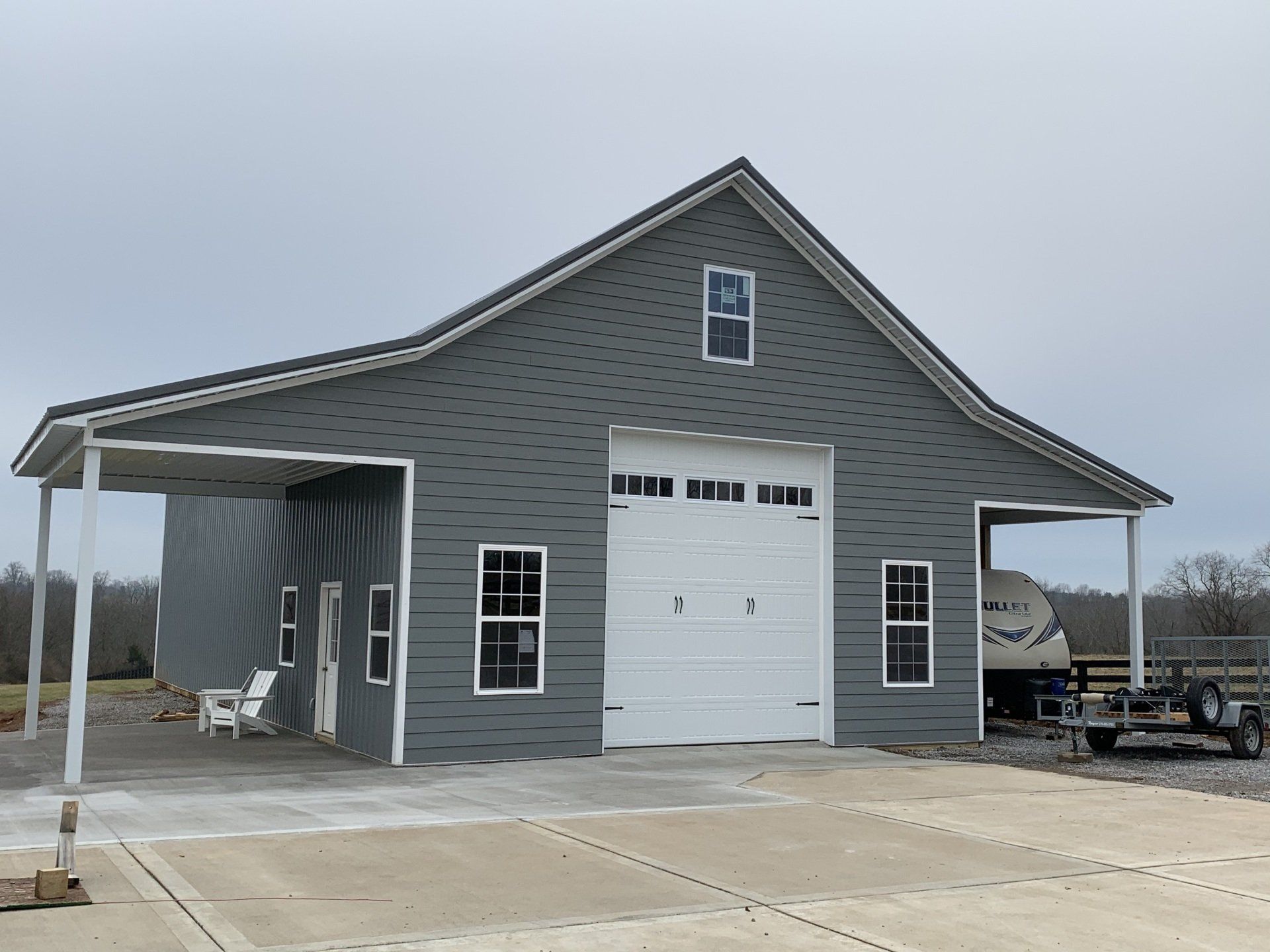 A large, gray barn-style building with a white garage door, two carports, and several windows on a concrete pad under an overcast sky.