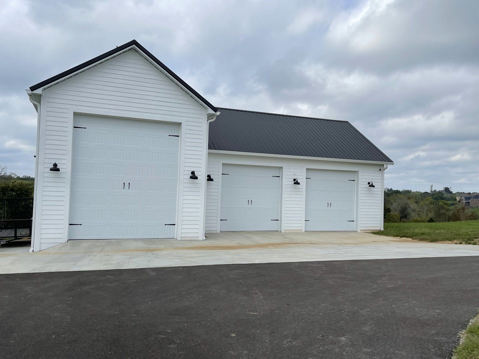 White garage with three bays, black roof, set against a cloudy sky, and asphalt driveway in the foreground.