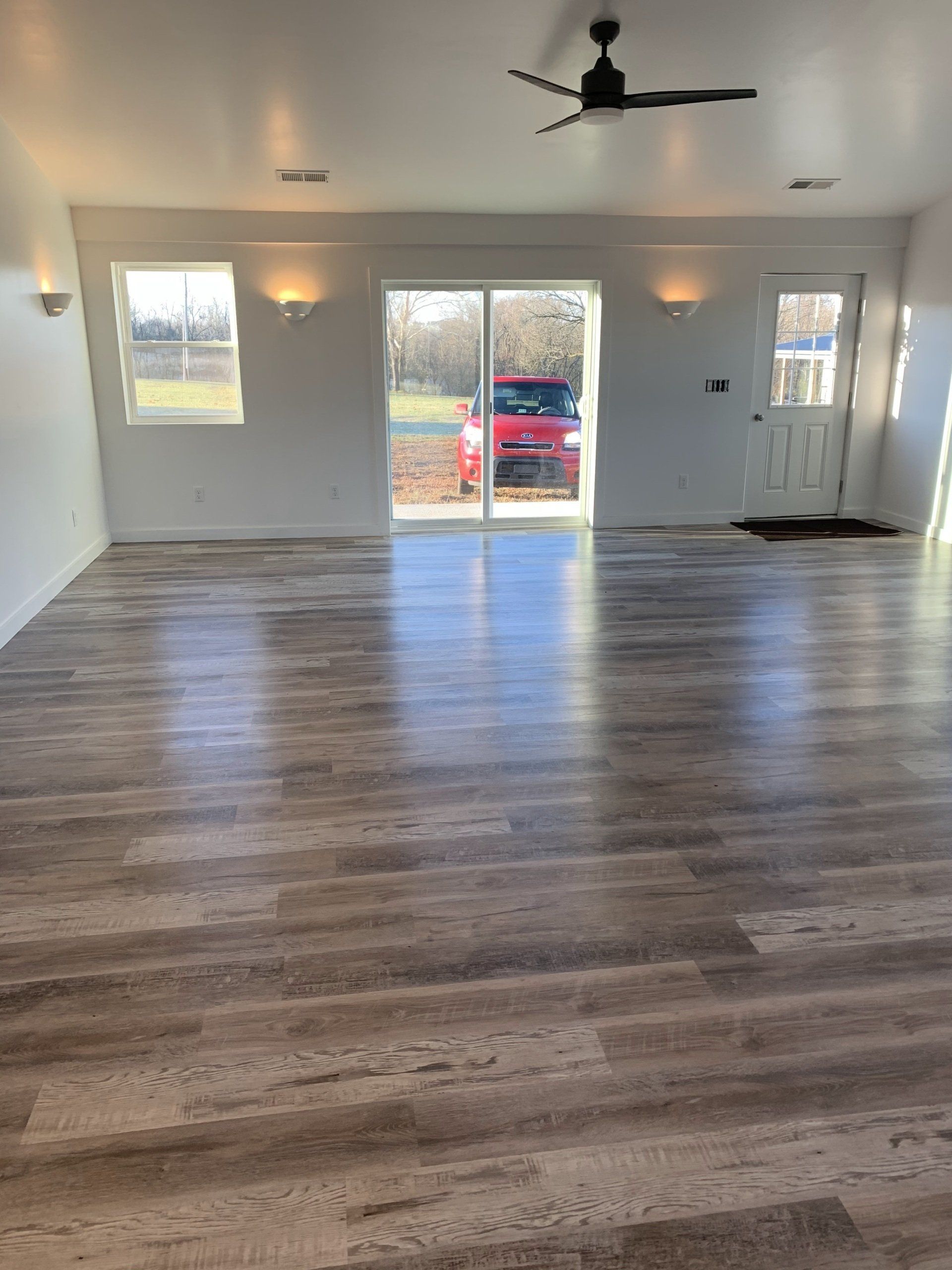 Empty room with light wood-look flooring and white walls. Sliding glass doors lead to a view of a red car and outdoor space.