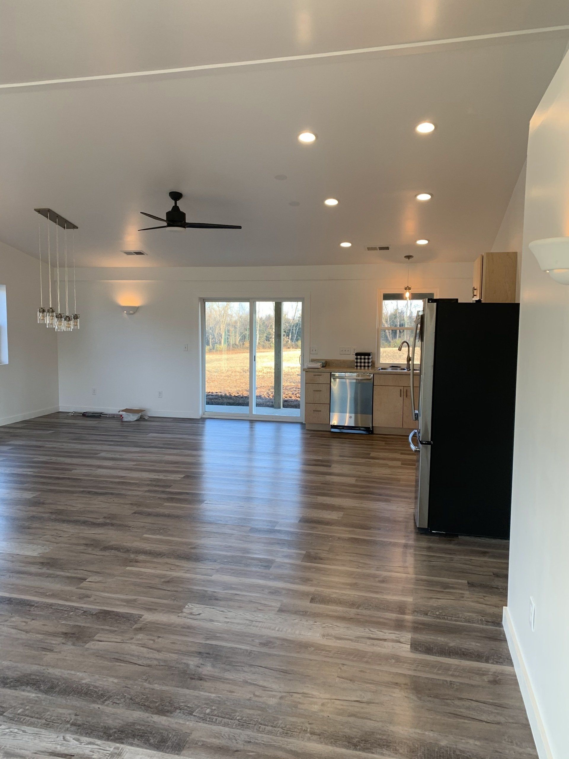 Spacious, modern living space with wood-look flooring, white walls, and recessed lighting. Includes kitchen area with stainless steel appliances and a black refrigerator.