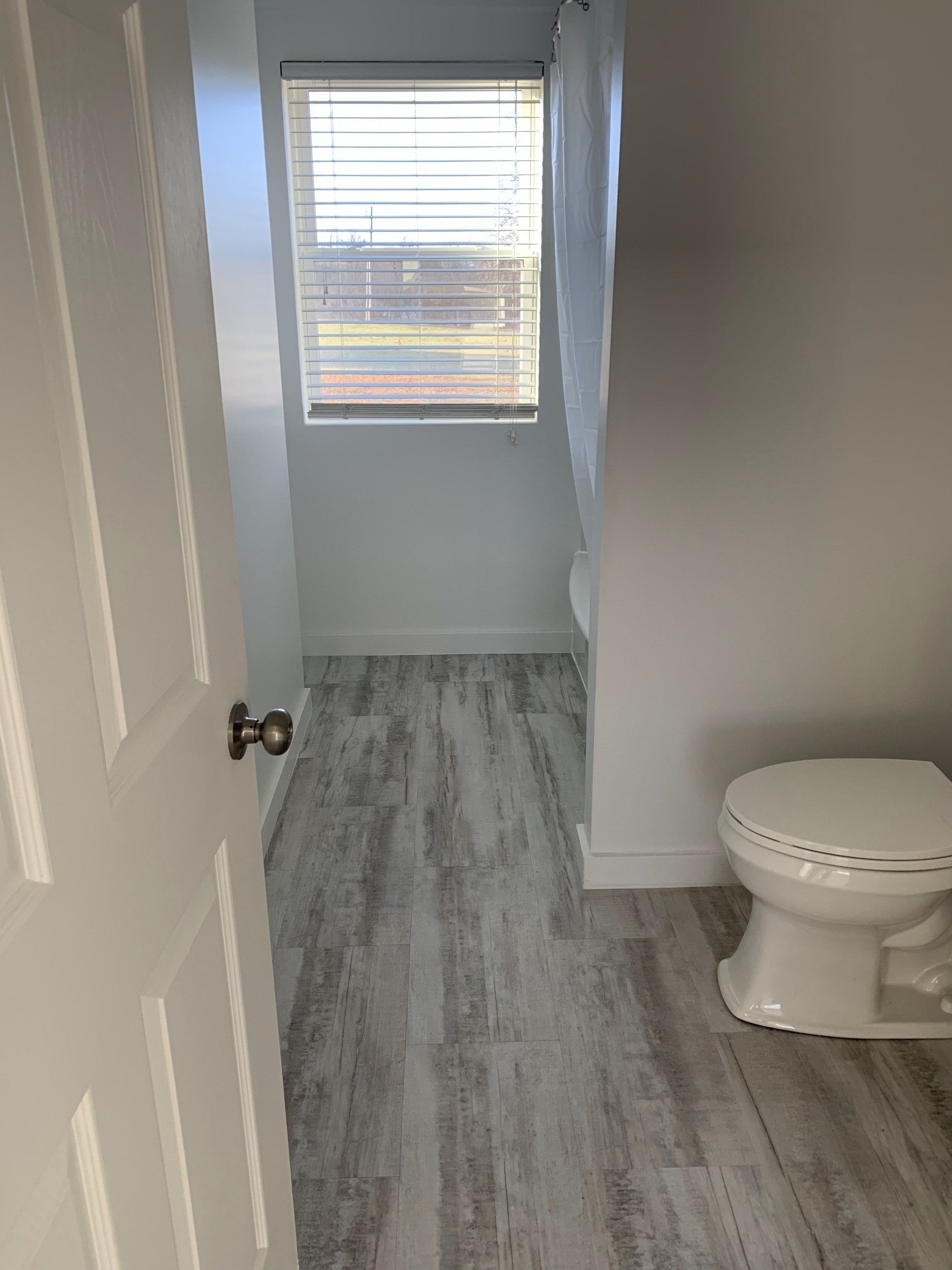 Bathroom with a white door, toilet, and window with blinds. Gray wood-look flooring and white walls.