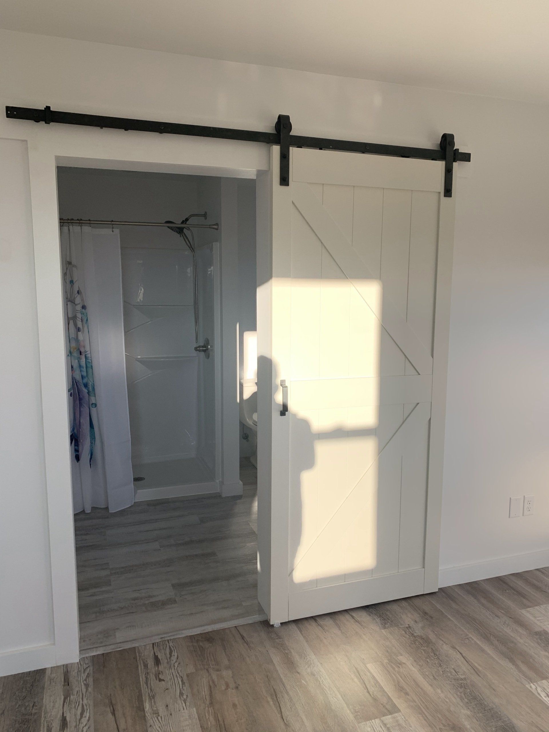 White barn door partially open to a bathroom, showing a shower and a light-colored floor. The door has a black sliding track.