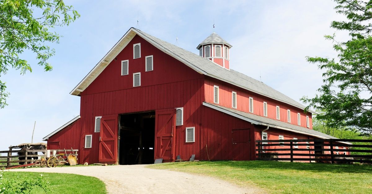 A large red pole barn stands on a grassy slope with a dirt path leading to its open doors on a brigh