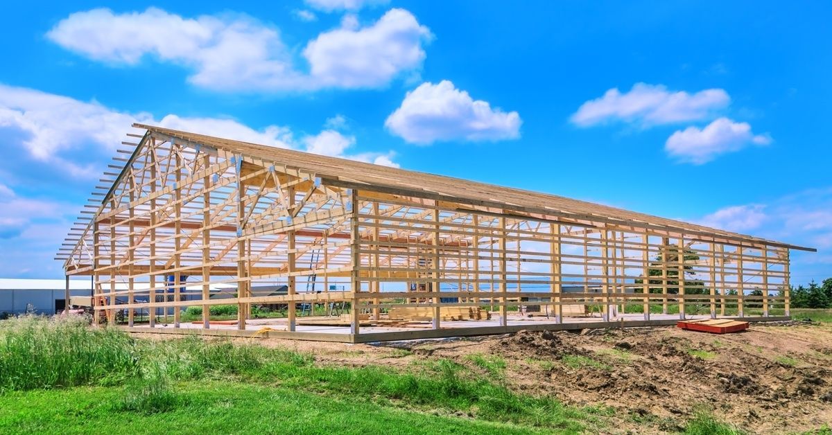A wooden pole barn frame under construction stands on a foundation, with open beams visible against a bright blue sky.