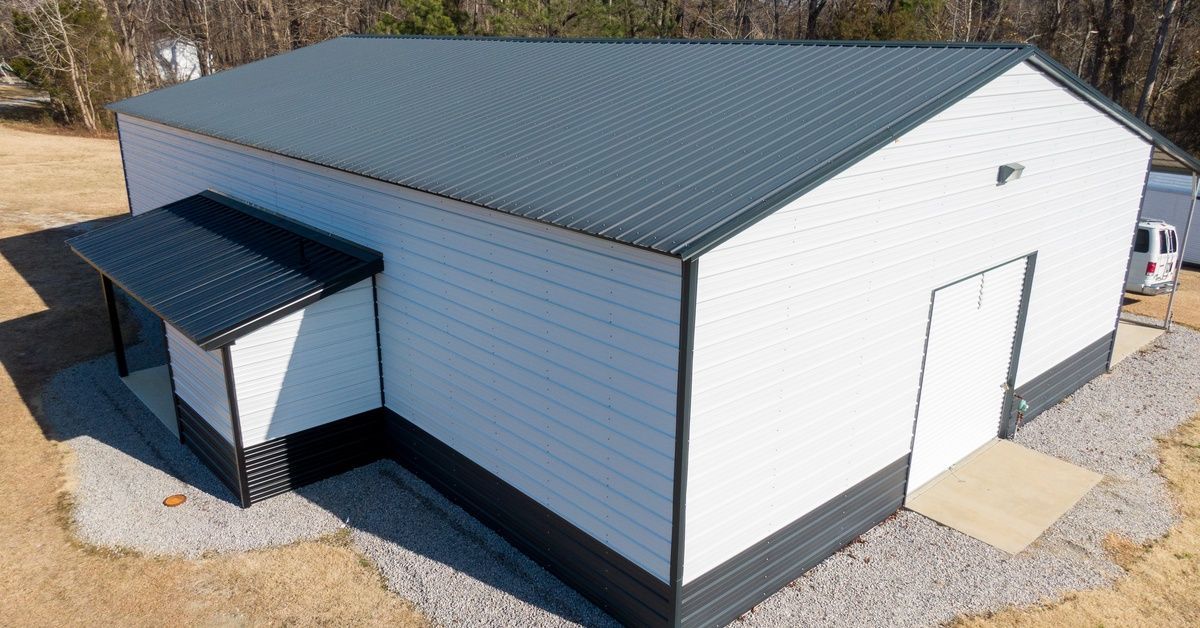 A newly built white pole barn with a metal roof sits on a gravel lot, surrounded by trees and dry gr