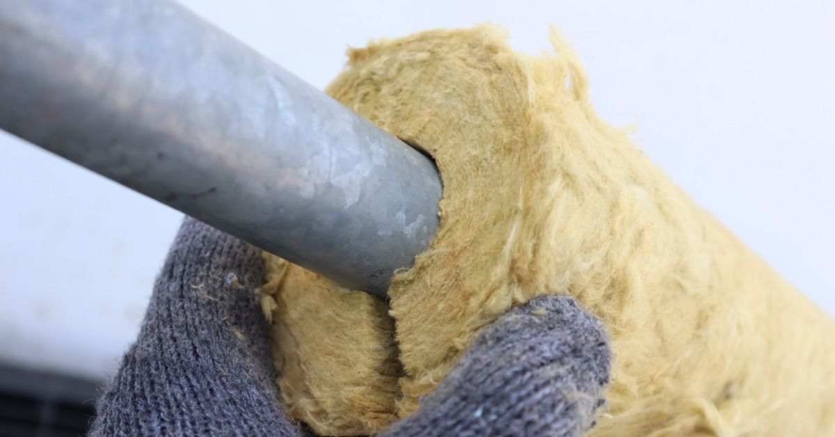 A close-up of a hand wearing a gray glove touching a roll of fiberglass insulation attached to a metal rod.