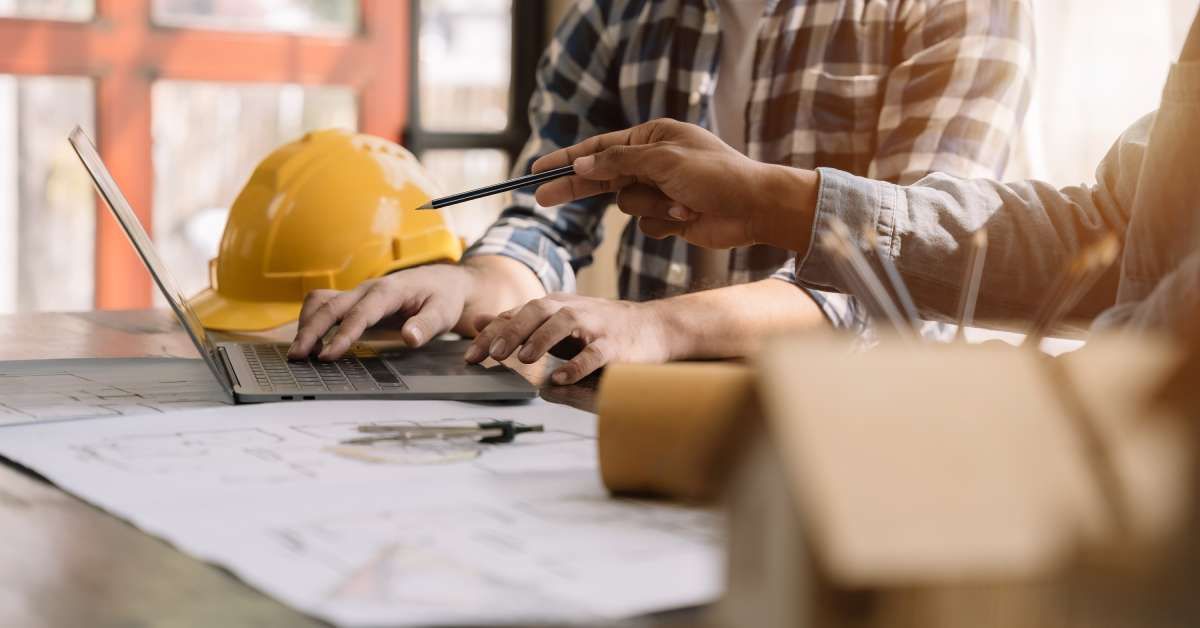 Two people sitting at a table, with a laptop and building plans in front of them. A yellow hard hat sits on the table.