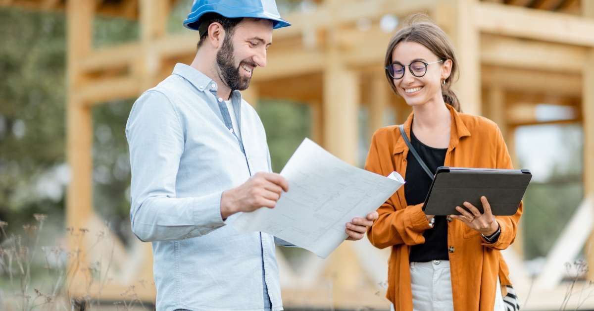 A man in a blue hard hat showing building plans to a smiling woman wearing glasses. They are outside near a construction site.