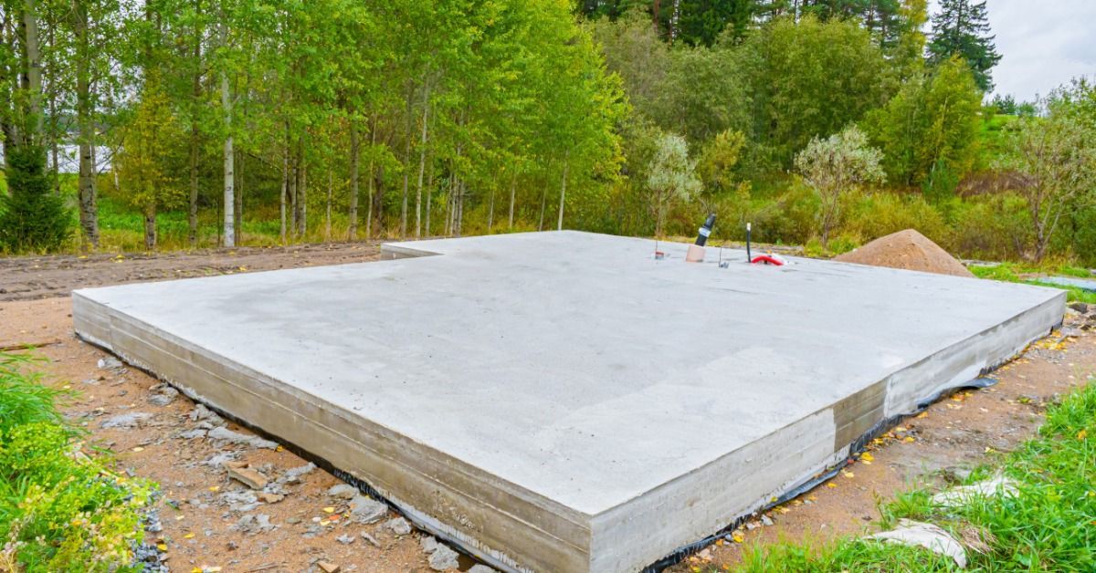 A concrete foundation slab surrounded by soil and trees at a construction site near a forested area.