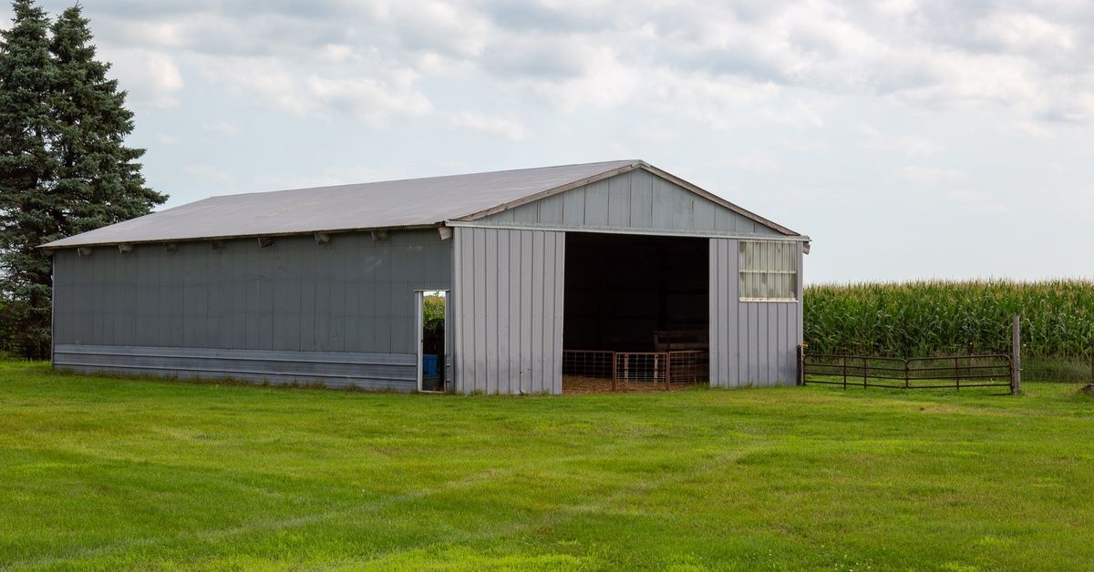 A gray metal pole barn with an open sliding door, mowed grass, and a cornfield beneath a cloudy sky.