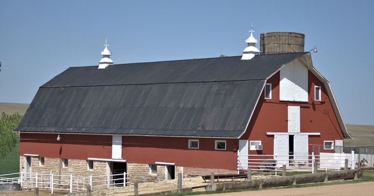 A red barn with white doors, a dark roof, fencing, a dirt road, and grassy hills under a clear blue 
