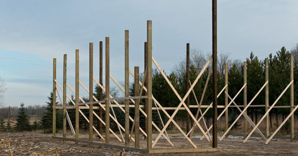 A wooden pole barn frame under construction with tall posts, diagonal braces, bare ground, and trees nearby.
