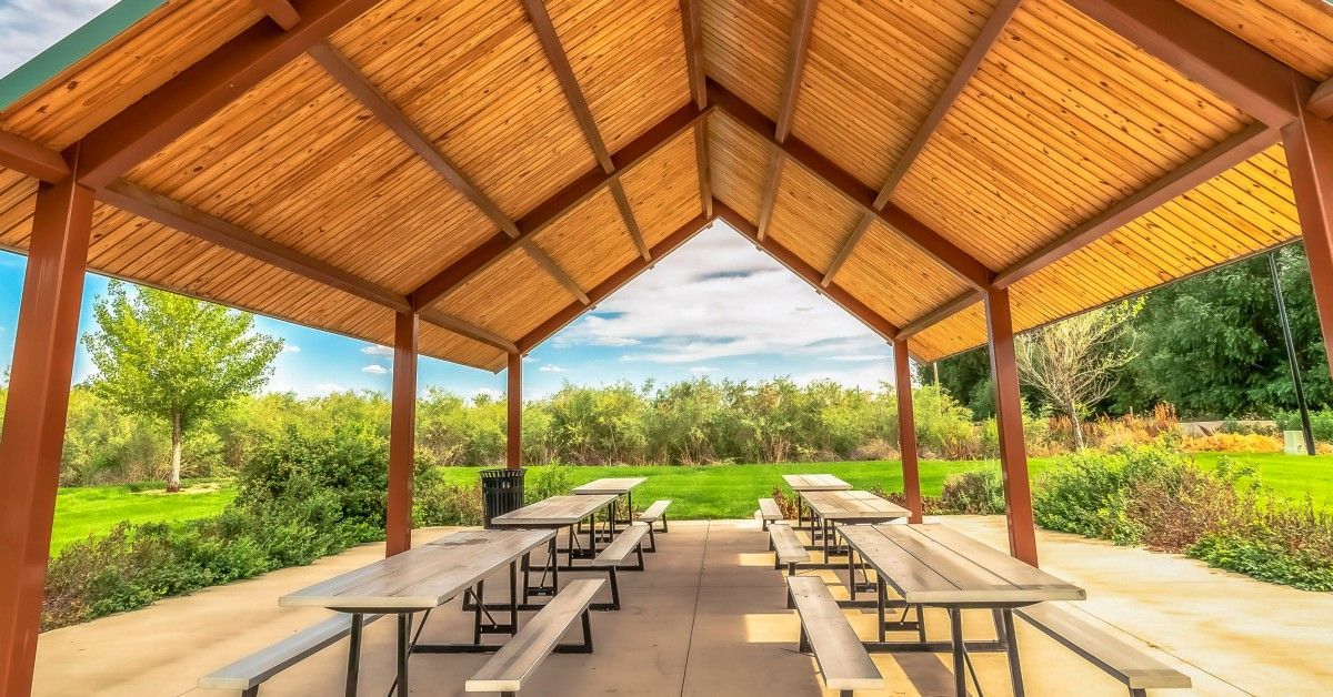  A view of a pole barn pavilion made out of wood. Six picnic tables are set up underneath on a concrete base.