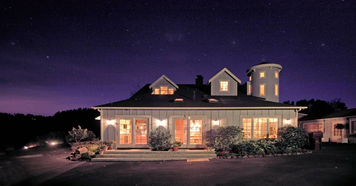 A white barn house lit up with warm indoor and outdoor lighting. Above the house is a blue-black sta