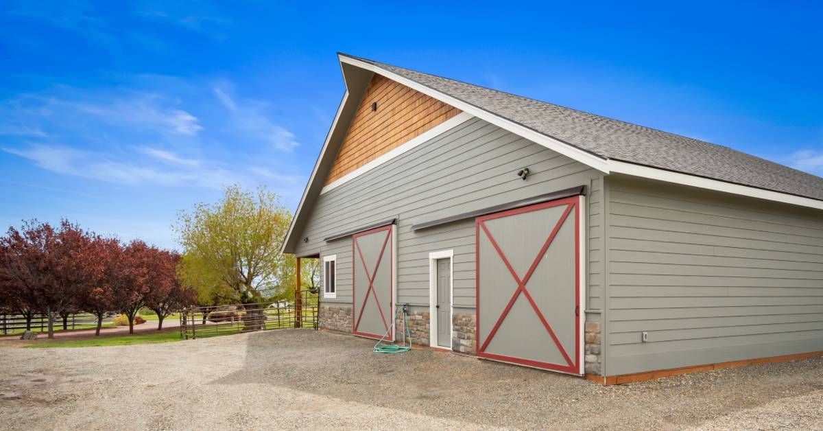 The exterior of a gray barn with red-accented doors and a stone border on the front-facing side. There are trees next to the barn.