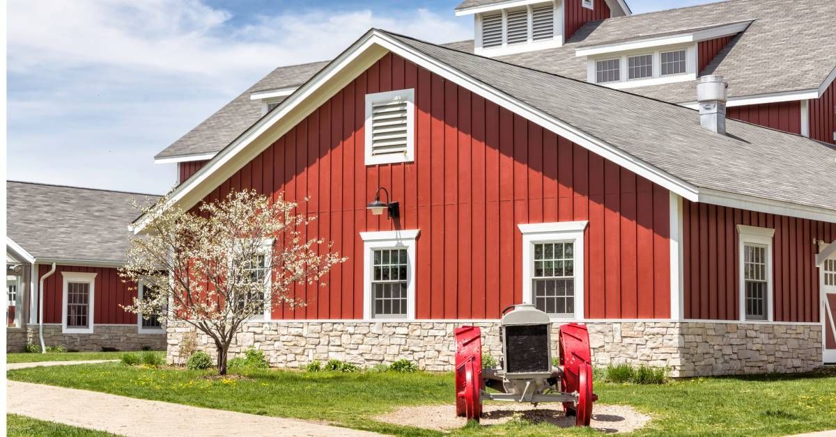 A red farmhouse with white windows and a stone border around the foundation. There is a small tractor on the front lawn.