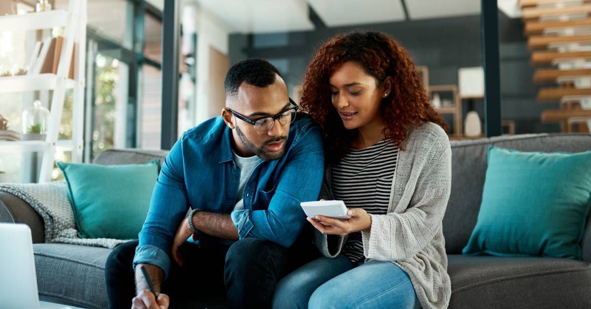 A couple sits on a gray couch, the woman holding a calculator and the man writing down numbers on a nearby form.