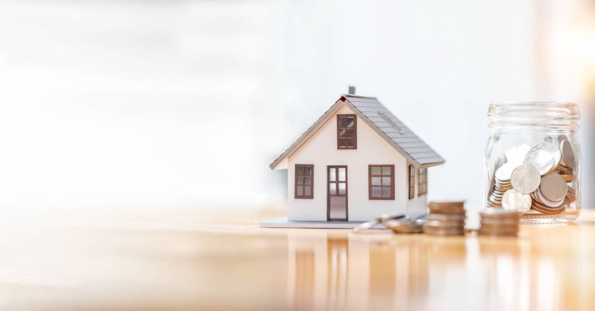 A miniature of a home sits on a table next to a glass jar full of coins, with several coins piled on