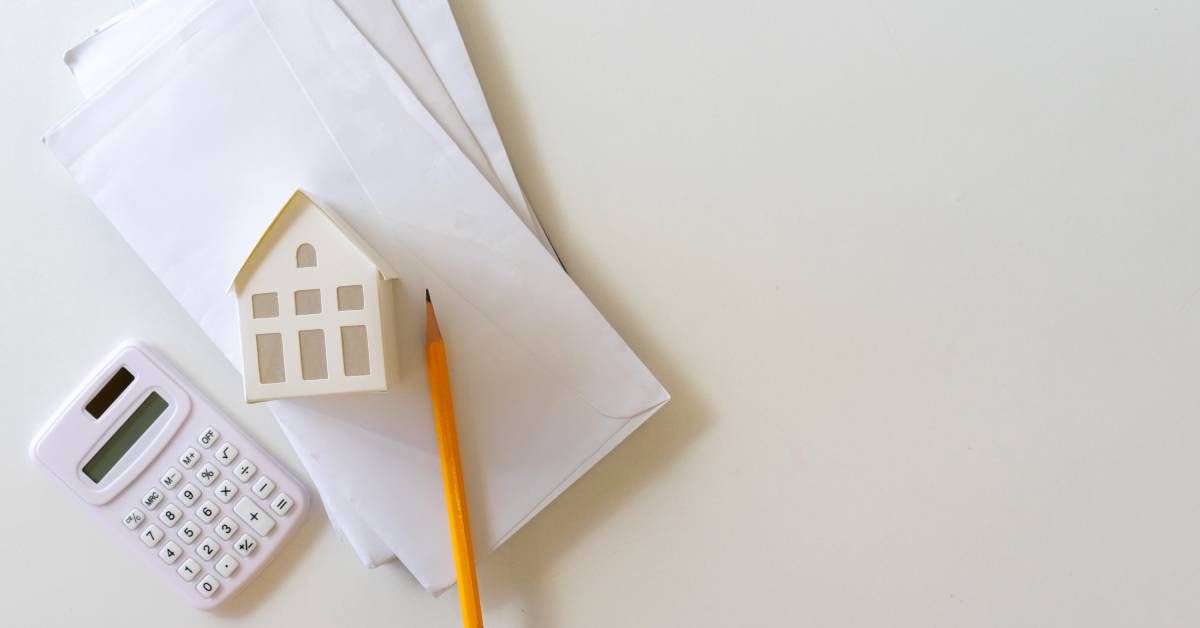 A miniature home rests on its side on top of a pile of white envelopes next to a calculator and pencil.