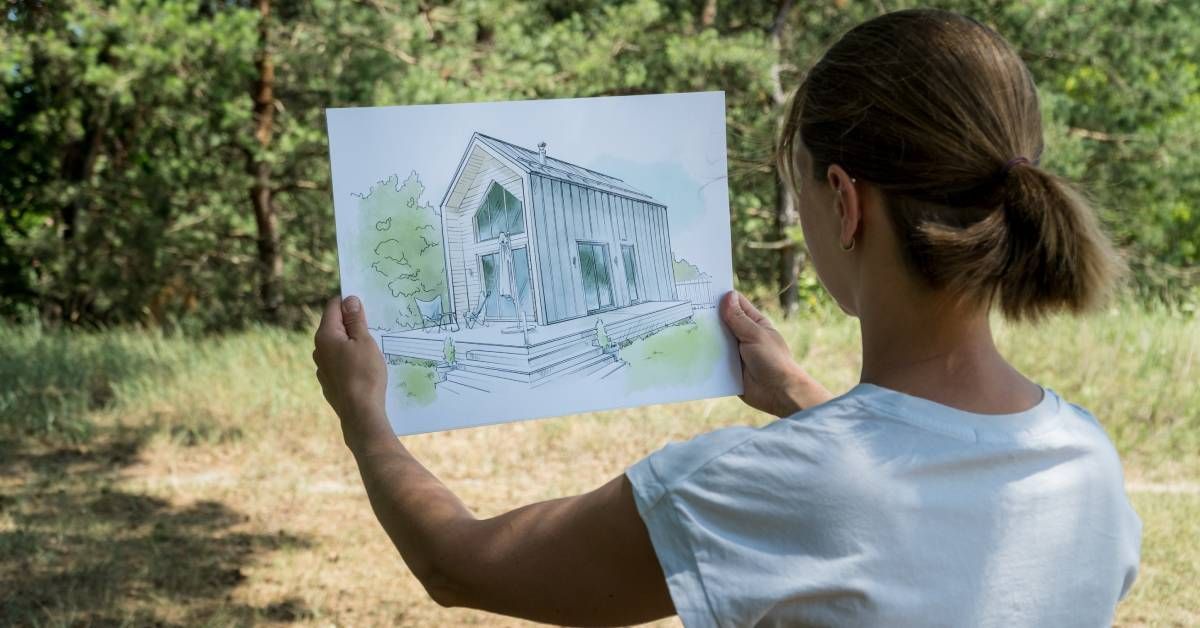 A woman holding an illustration of a barn home and comparing it with the land in front of her. She stands in the shade.