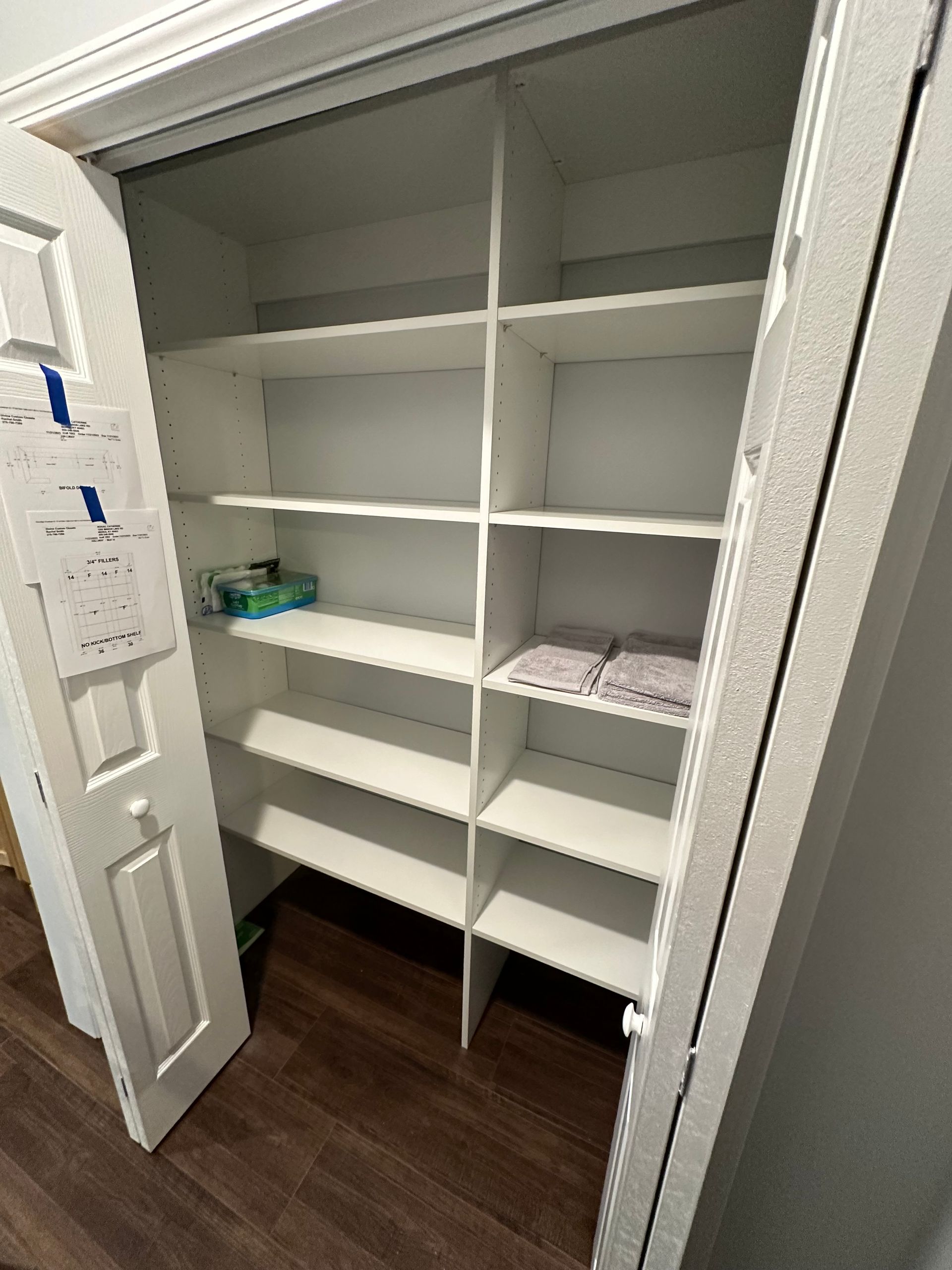 White, empty closet with built-in shelves. Partially open white door on the left, with a wood floor.