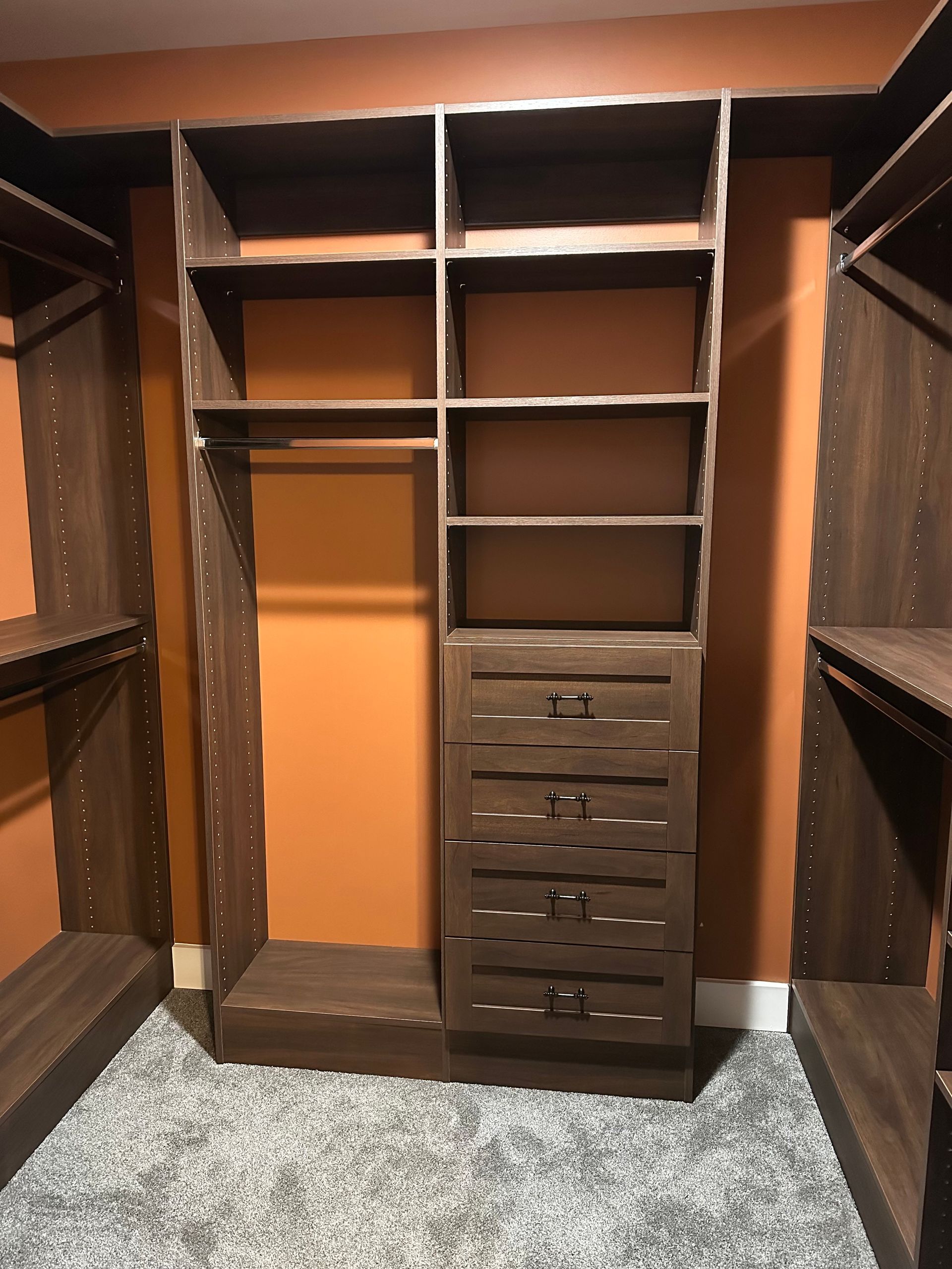 Dark wood closet organizers in a room with orange walls and gray carpet. Central unit has shelves, hanging rod, and drawers.