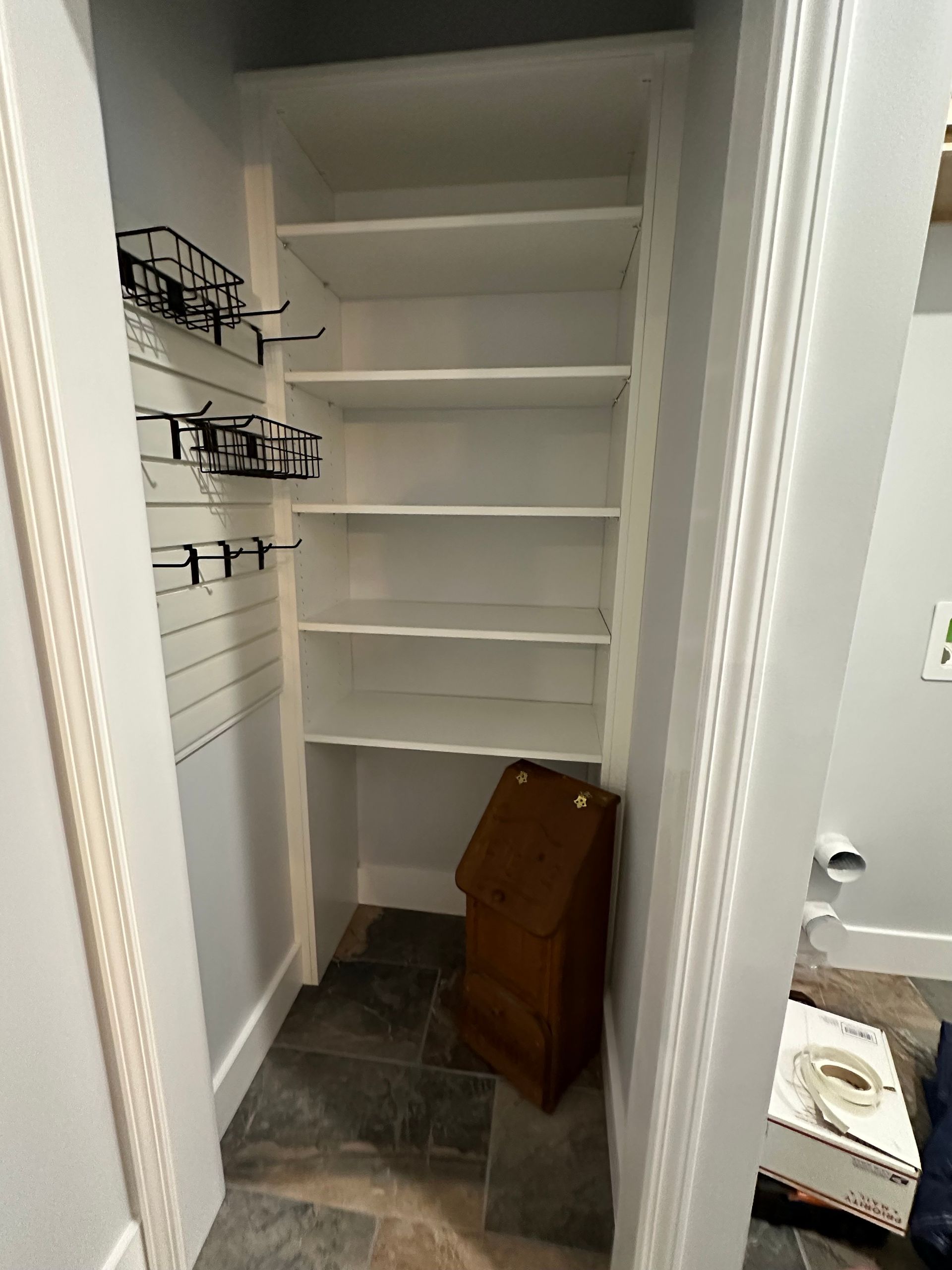 Built-in white shelving in a narrow closet space, with a brown wooden box at the bottom and wire storage racks on the left wall.