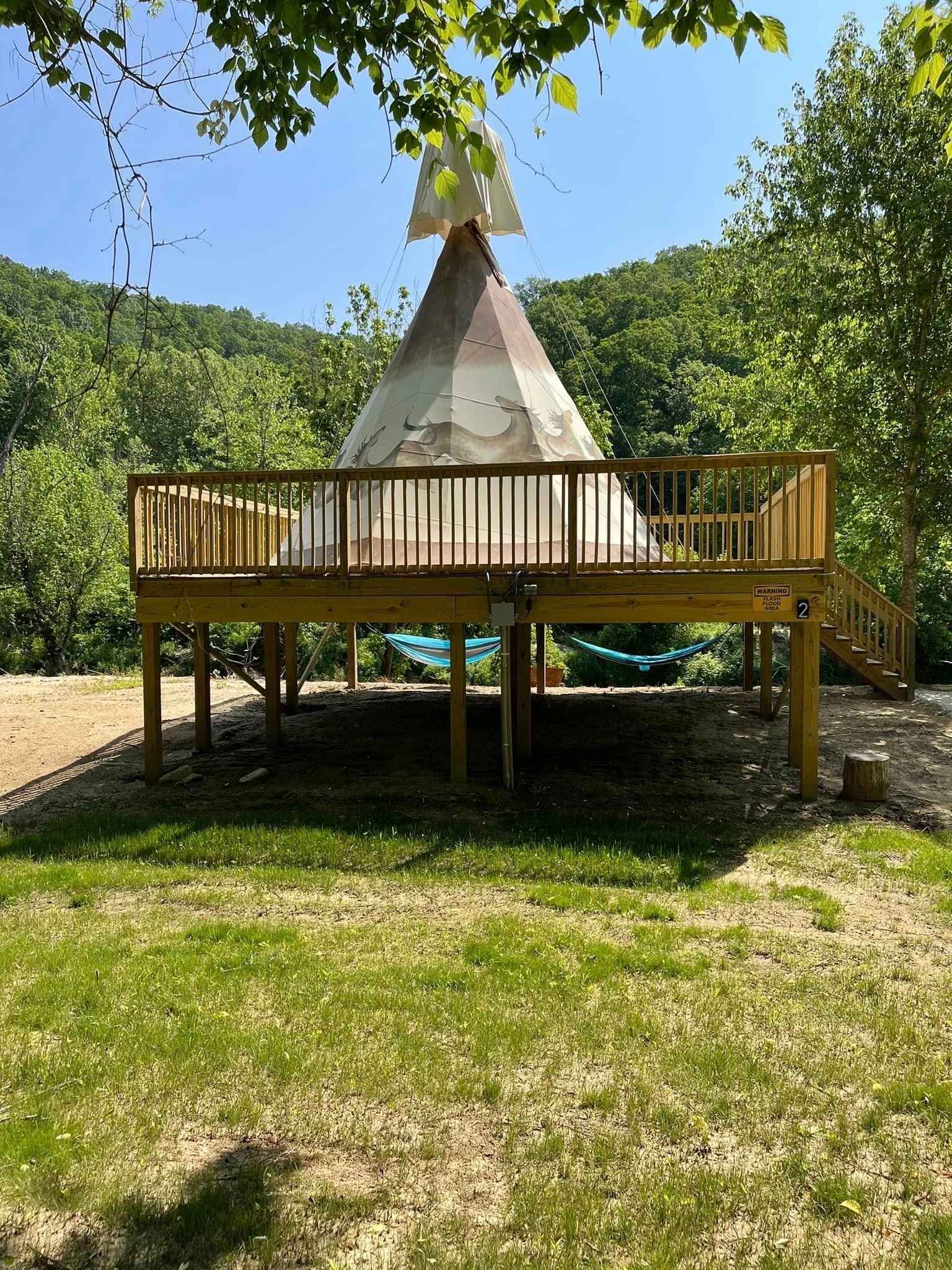 A raised wooden deck with a teepee-like structure on it, in a wooded outdoor setting.