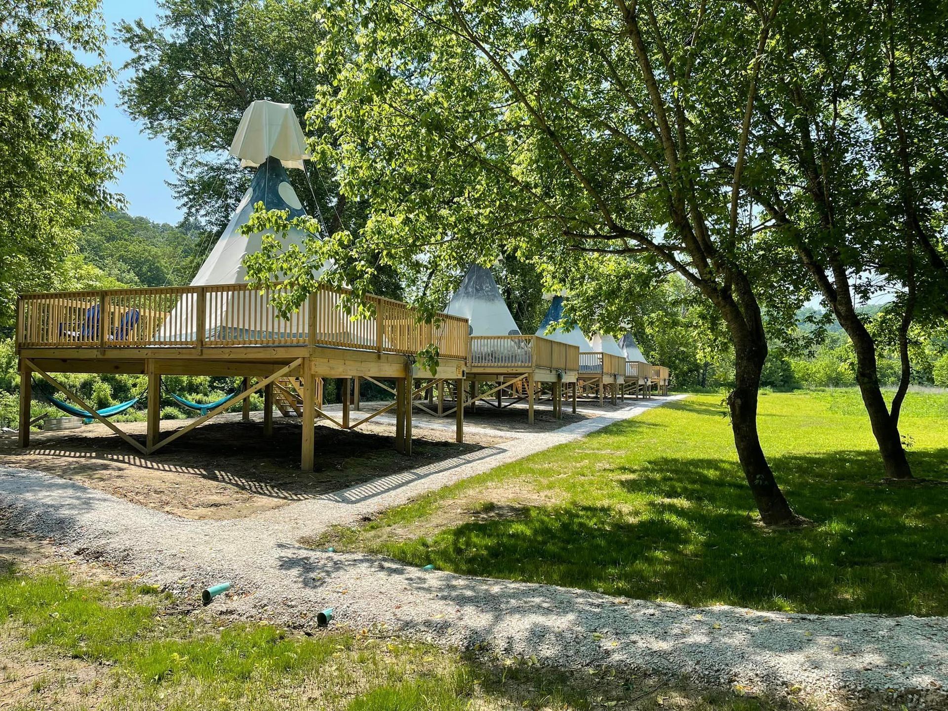 Row of teepee-style structures with wooden decks in a green, tree-filled outdoor setting.